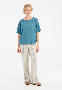 Mėlynos žalios chevron raštų laisva palaidinė, derinama su dryžuotomis, šviesių spalvų plačiakelnėmis kelnėmis, turinčiomis geltonų ir mėlynų akcentų. Patogi avalynė.