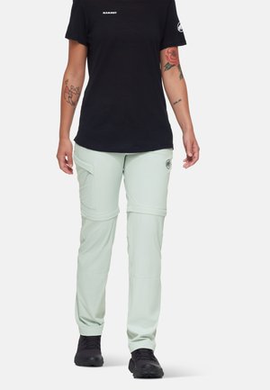 Person trägt hellgrüne Zip-off-Wanderhose, ein schwarzes Mammut-T-Shirt, schwarze Schuhe und sichtbare bunte Arm-Tattoos.