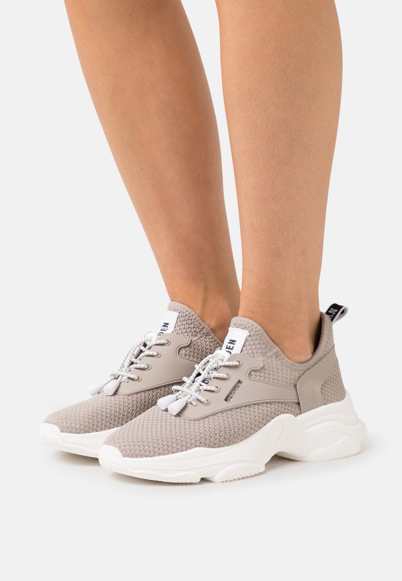 Baskets tricotées beige clair avec semelles épaisses blanches et lacets élastiques à motifs, portées pieds nus sur fond blanc.