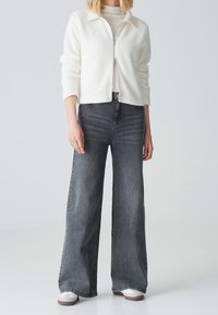 Mujer con chaqueta de forro polar blanca con cremallera, top beige claro, vaqueros negros desgastados de pierna ancha y zapatillas blancas, de pie contra un fondo liso.