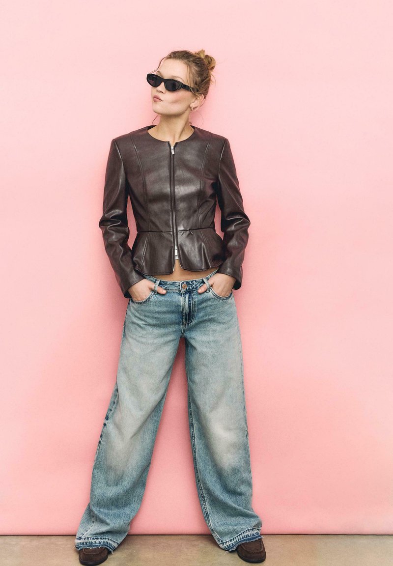 Jeune femme portant des lunettes de soleil noires, une veste en cuir marron et un jean bleu à jambes larges, debout les mains dans les poches devant un fond rose.