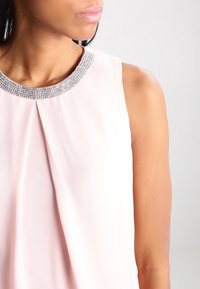 Blusa sin mangas de color rosa con un frente plisado y un escote de cuentas plateadas brillantes, con una textura de tela suave y ligera.