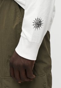 Λευκό πουκάμισο με μακρύ μανίκι και μαύρο σχέδιο ιστού αράχνης στο αριστερό μανίκι, σε συνδυασμό με χακί πρασινωπό σαλοπέτα. Λείος βαμβακερός υφασμάτινος ιστός.