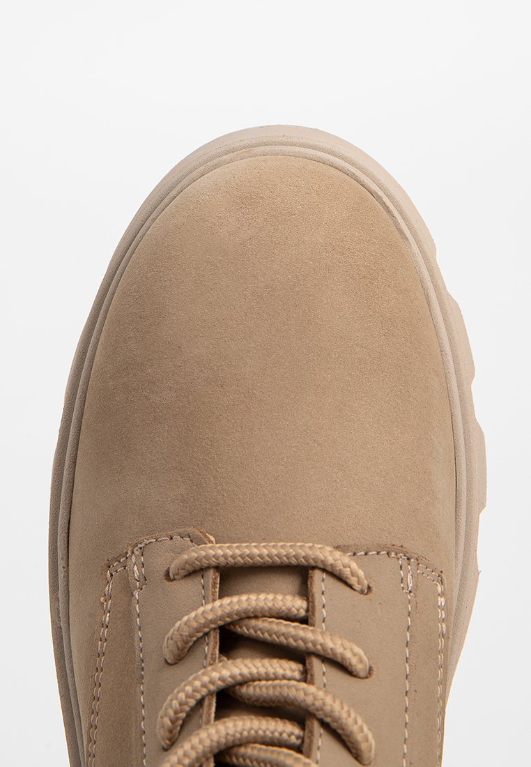 Beige Wildlederschuh mit runder Zehenform, strukturiertem Obermaterial und kontrastierenden Nähten. Verfügt über geflochtene Schnürsenkel und eine dicke Gummisohle.