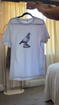 Valge puuvillane T-särk ümmarguse kaelusega, millel on graafika tuvist, kes seisab sneakeritel. Lühikesed varrukad ja lõdva lõikega disain.