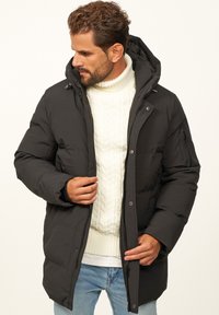 Schwarze Steppjacke mit Kapuze, Reißverschluss und Druckknopfverschluss, seitlichen Taschen und einem strukturierten weißen Pullover darunter. Lässige Jeans.