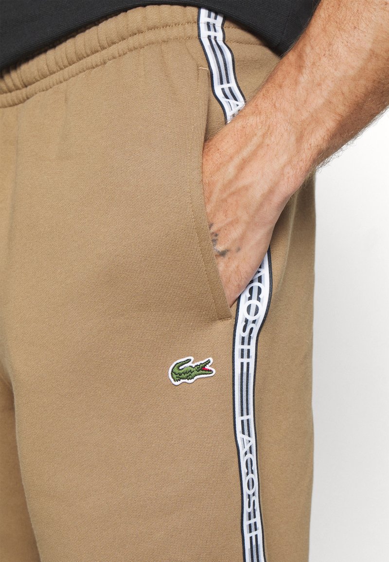 Pantalons de survêtement beiges avec une coupe slim, dotés de poches latérales, d'accents à rayures blanches et d'un logo crocodile brodé en vert sur la cuisse gauche.