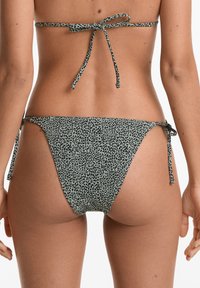 Bikini-Unterteil mit Tiermuster in hellgrün mit schwarzen Punkten. Schnüre an jeder Seite, figurbetontes Design und glatte Textur.