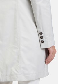 Lichtgrijze jas gemaakt van gladde stof, met een rechte snit, vier knopen op de mouwen en subtiele stikdetails langs de naden.