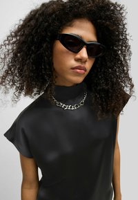 Schwarzes Satinoberteil mit kurzen Ärmeln, massiver silberner Kettenhalskette und eckigen schwarzen Sonnenbrille. Lockige Haare umrahmen das Gesicht.