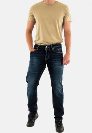 Beige crew-neck t-shirt paired with dark blue denim jeans. Jeans feature classic five-pocket design and subtle stitching accents. Black sneakers.