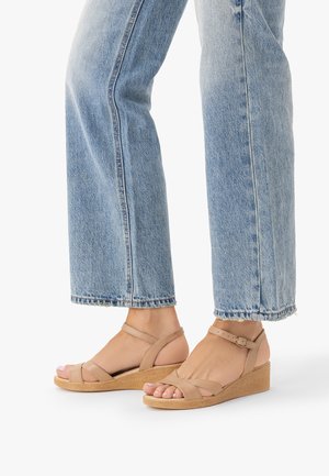 Stopy w beżowych sandałach na koturnie z paskami wokół kostki oraz jasnoniebieskich dżinsach, na białym tle.