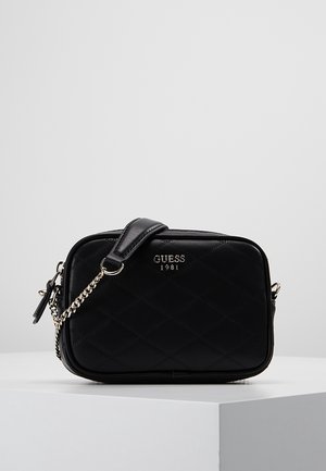 Schwarze gesteppte Leder-Guess-Crossbody-Tasche mit silberner Kettenriemen und Logo vorne, auf einer weißen Oberfläche vor einem hellgrauen Hintergrund platziert.