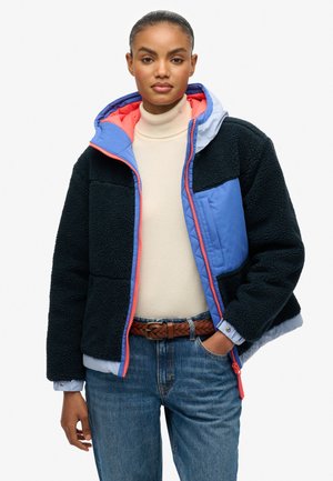 Schwarze Sherpa-Jacke mit hellblauen Akzenten und korallenfarbigem Reißverschluss. Verfügt über eine Kapuze und eine gesteppten Brusttasche. Kombiniert mit einem cremfarbenen Rollkragenpullover.