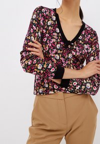 Pull en maille à motif floral noir avec des motifs roses, jaunes et blancs, doté d'un col en V et de poignets côtelés. Associé à un pantalon beige.