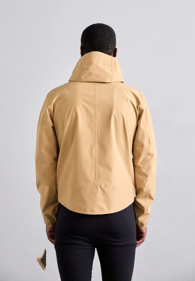 Veste légère beige avec un col montant, présentant une texture lisse et un ourlet arrondi. Portée avec des leggings noirs texturés.