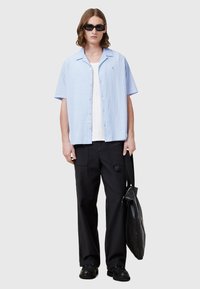 Lichtblauw kortom-shirt met een gestructureerde afwerking, zwarte broek, zwarte sneakers en een zonnebril. Draagt een zwarte schoudertas.