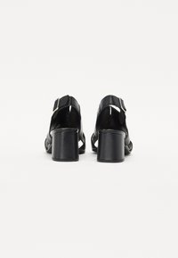 Talons en vernis noir avec un talon épais, une sangle réglable, un design découpé et une texture lisse. Vue arrière mettant en valeur la hauteur du talon.