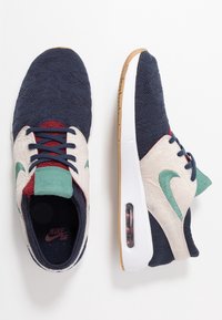 Baskets bleu marine et crème avec un design texturé, comprenant des accents verts et rouges, une semelle blanche, et une unité d'air visible au talon.