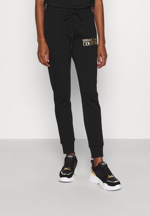 Personne portant un jogging noir Versace Jeans Couture avec logo doré et des baskets épaisses noires, blanches et dorées, debout devant un fond uni.