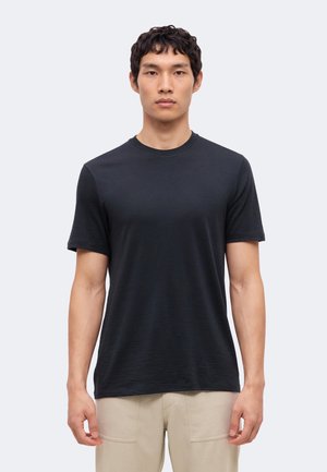 Jeune homme portant un t-shirt noir uni à manches courtes et un pantalon beige clair, debout devant un fond clair uni.