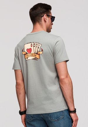 Homme portant un t-shirt gris avec des cartes à jouer (As, Roi, Reine, Valet) et une bannière "FORTUNE" imprimée dans le dos, associé à un jean bleu.