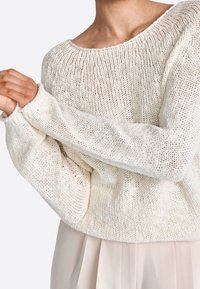 Suéter de punto beige claro con un corte holgado, escote ancho y mangas largas. La textura es abierta y aireada, con un patrón de canalé.