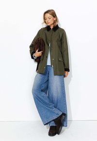 Veste imperméable vert olive avec un col noir, associée à un jean en denim bleu ample et des bottines en daim marron. Tenant un sac à franges marron.