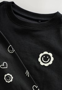 Sweat-shirt noir avec un col côtelé orné de motifs imprimés blancs : visages souriants, cœurs et une fleur. Texture douce et design décontracté.