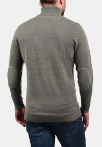 Pull en laine grise avec un col haut, des bordures côtelées sur l'ourlet et les poignets, doté de patchs au niveau des coudes et d'une coupe ajustée.