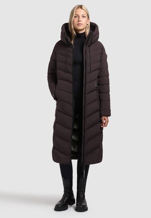 Kvinna iklädd en lång svart vadderad kappa med huva, svart polotröja, svarta tights och svarta stövlar som når till vaden, stående mot en enfärgad bakgrund.