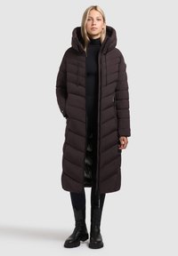 Vrouw in een lange zwarte pufferjas met capuchon, zwarte coltrui, zwarte leggings en zwarte laarzen tot halverwege de kuit, staande tegen een effen achtergrond.