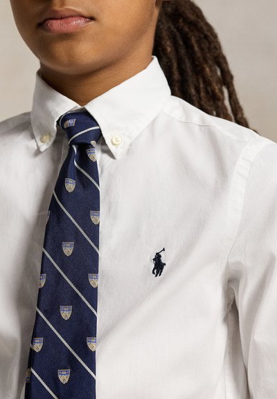 Personne portant une chemise blanche boutonnée avec une cravate rayée bleu marine présentant des motifs de blasons jaunes et bleus.