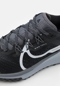 Gros plan sur une chaussure de course Nike Trail noire et grise avec des lacets texturés et un Swoosh blanc sur une semelle grise.