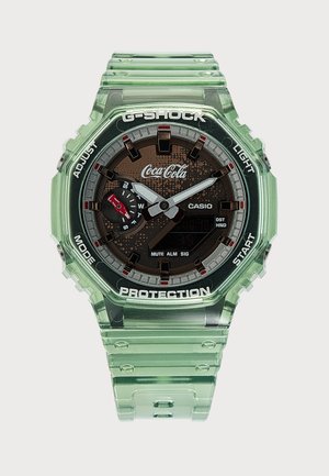 Διαφανές πράσινο ρολόι Casio G-Shock με λογότυπο Coca-Cola, αναλογική και ψηφιακή οθόνη και ετικετοποιημένα κουμπιά στο στεφάνι.