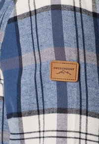 Tissu à carreaux bleu et blanc en gros plan avec un patch rectangulaire beige marqué "Swedemount" et un logo de montagne sous le texte.