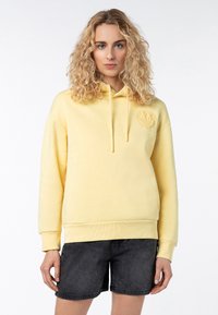 Geel hoodie met een voorzak, ribgebreide manchetten en een gestructureerd logodetail op de borst. Gecombineerd met zwarte shorts.