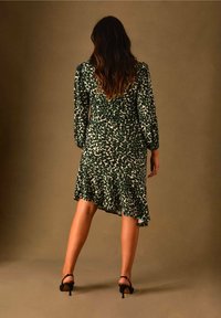 Zelené a bílé šaty s leopardím potiskem, dlouhými rukávy a volánkovým lemem. Nošeny s černými sandály na podpatku s páskem. Texturovaná tkanina s přiléhavým střihem.