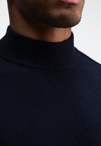 Navyblauer Rollkragenpullover mit geripptem Kragen und glatter Textur. Eine Nahaufnahme hebt das gestrickte Material und das Detail des Ausschnitts hervor.