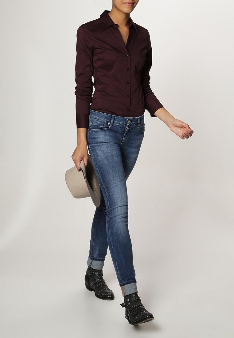 Camisa burdeos de botones, ajustada, de manga larga. Jeans de mezclilla azul, de corte slim, con los puños doblados en los tobillos. Botas de tobillo negras con tachuelas, sombrero gris en la mano.