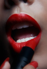 Se aplica pintalabios rojo en unos labios carnosos, mostrando un acabado brillante. Los dientes son brillantes y blancos. El tubo de pintalabios es negro con una punta roja.