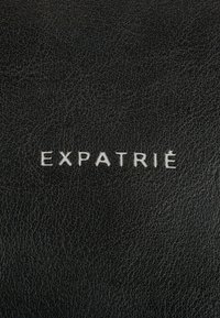 Black textured leather with embossed white text "EXPATRIÉ" prominently displayed at the center.