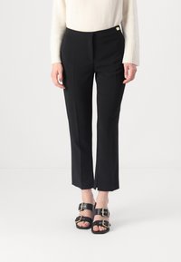 Pantaloni neri su misura con una leggera svasatura e pieghe frontali, dotati di hardware dorato in vita, abbinati a sandali neri.