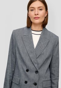 Grijze dubbelgeknoopte blazer van textuurstof met geknipte revers, twee voorkreidjes en zwarte knopen. Draagt over een gestreept shirt.