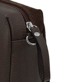 Acercamiento de la esquina de un bolso de cuero texturizado marrón oscuro con cremallera y correa de cuero unida, asegurada por un anillo de metal y remache.