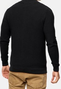 Mann trägt einen schwarzen gestrickten langärmeligen Pullover und tanfarbene Hosen, von hinten vor einem schlichten weißen Hintergrund gezeigt.