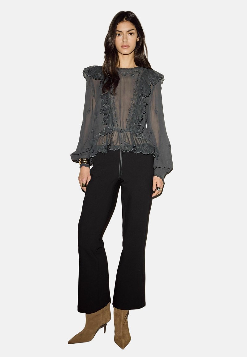 Kvinde iført en gennemsigtig mørkegrå bluse med flæser, sorte vide bukser, beige ankelstøvler med spids tå samt gyldne armbånd og ringe.