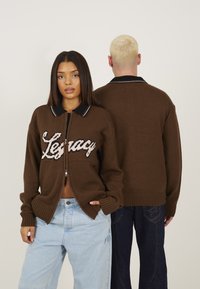 Pull en tricot brun avec col noir et rayure blanche. Présente le texte "Legacy" sur le devant du zip. Le modèle porte un jean clair.