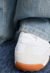 Zapatilla blanca con acentos de ante y cuero, suela de goma naranja y diseño de cordones, parcialmente cubierta por pantalones de mezclilla azul claro.