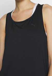 Czarny sportowy top na ramiączkach z teksturowanym wzorem, z okrągłym dekoltem i małym czarnym logo Puma z przodu. Noszony warstwowo z białą koszulką pod spodem.